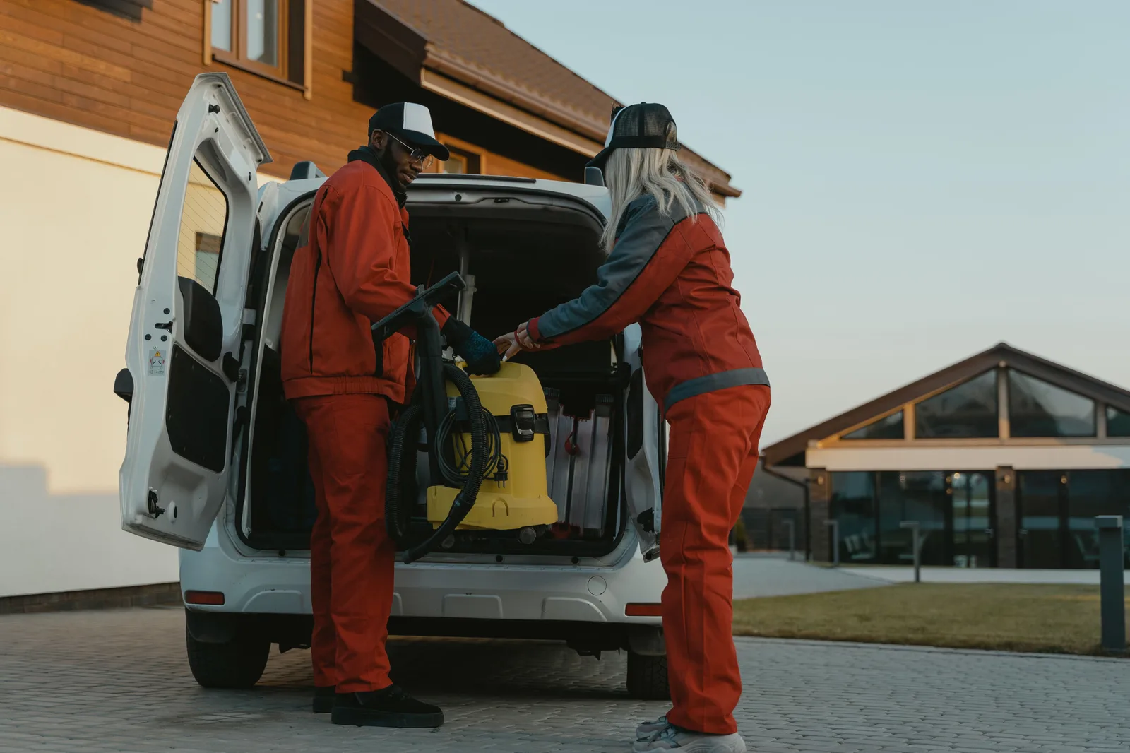 Two cleaners loading a vacuum into a van outside a modern building at dusk. Window cleaning referrals: four sources, seven steps, and the sentences that work