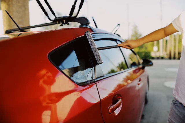 Close-up of a person using a squeegee to clean a red car window outdoors. Top profitable window cleaning services options compared and reviewed