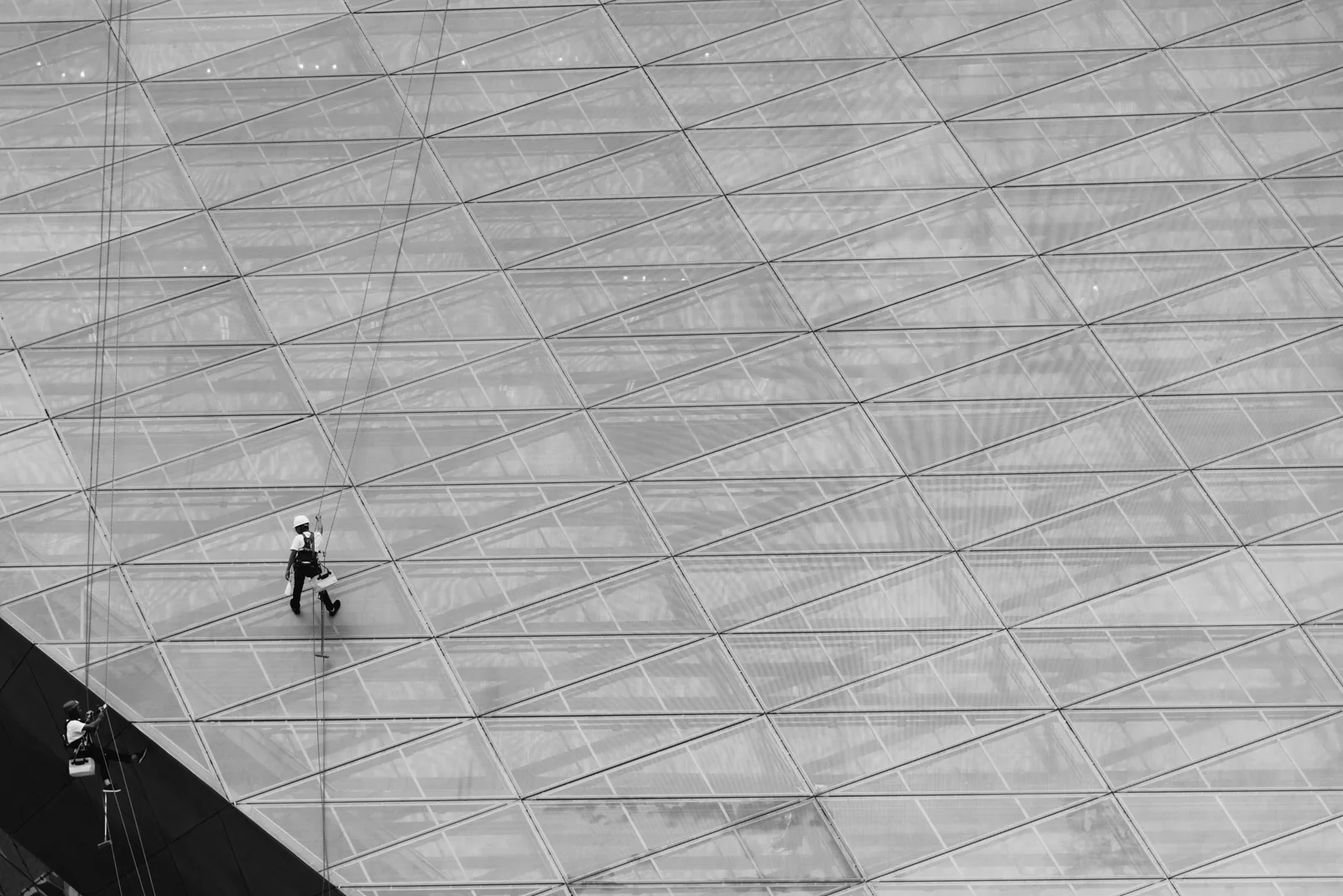 A monochrome image of workers cleaning a large glass structure using ropes. Window cleaning staffing: from hire to trained