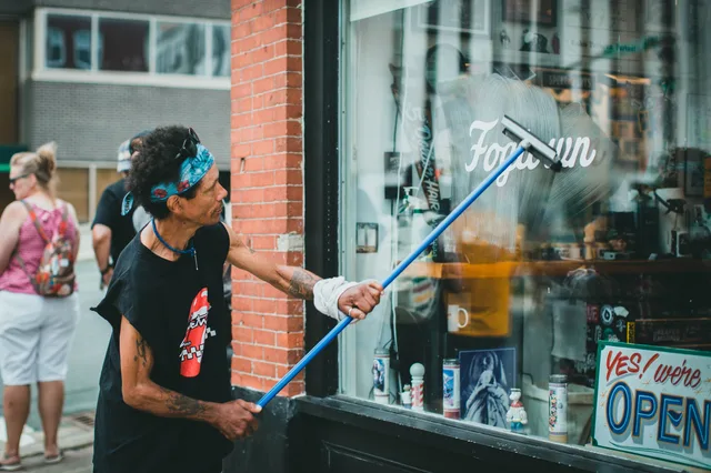 A man with tattoos cleaning a glass storefront window on a busy street using a squeegee. Seven window cleaning trends to check in your own market